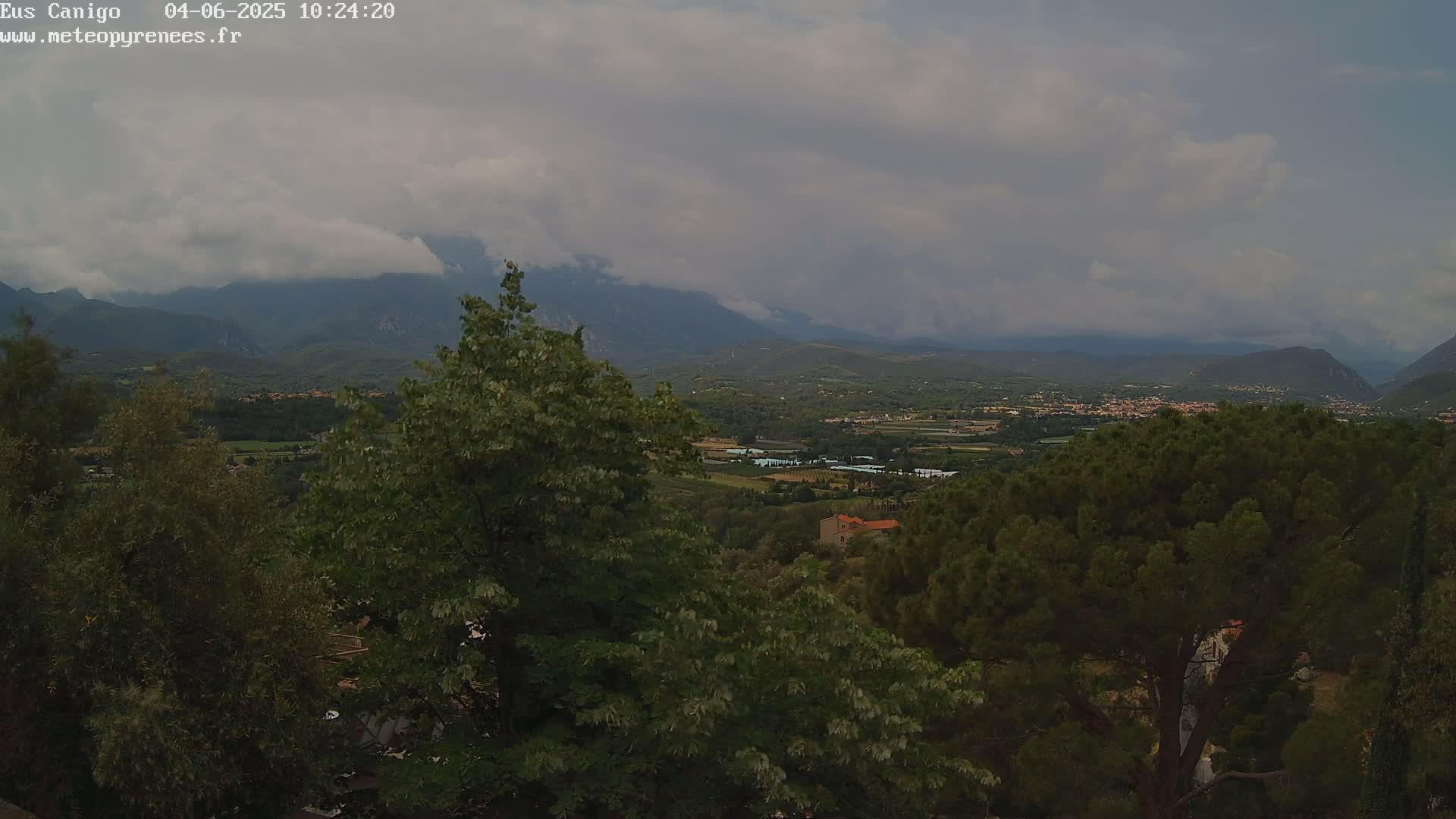 A partly cloudy sky overlooks a valley of green trees, shrubs, and distant buildings nestled between mountains.