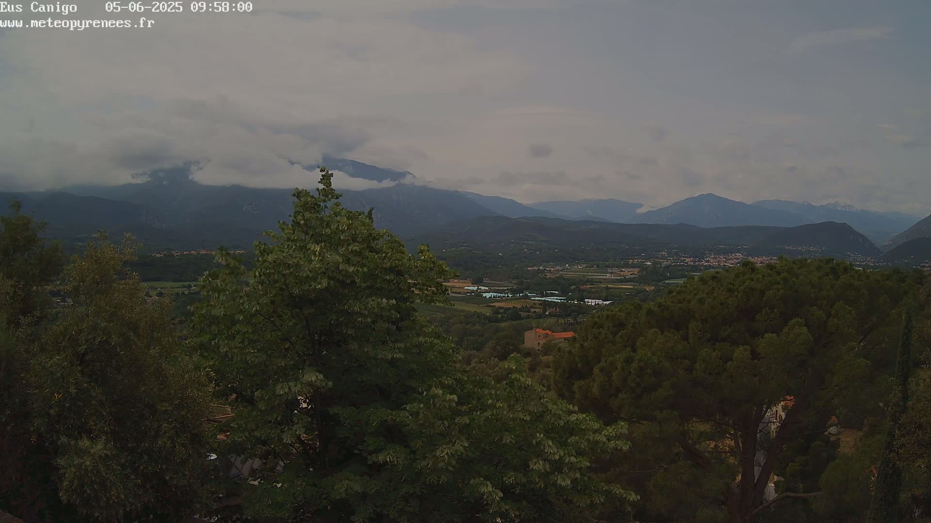 A view of a valley nestled between mountains under an overcast sky, partially obscured by lush green trees in the foreground.