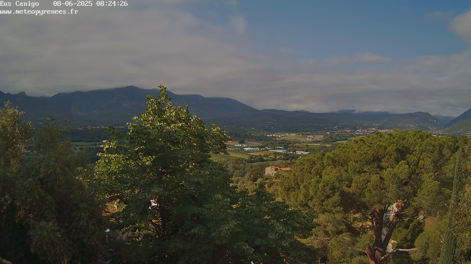 A partly cloudy sky overlooks a valley with green trees, buildings, and distant mountains.