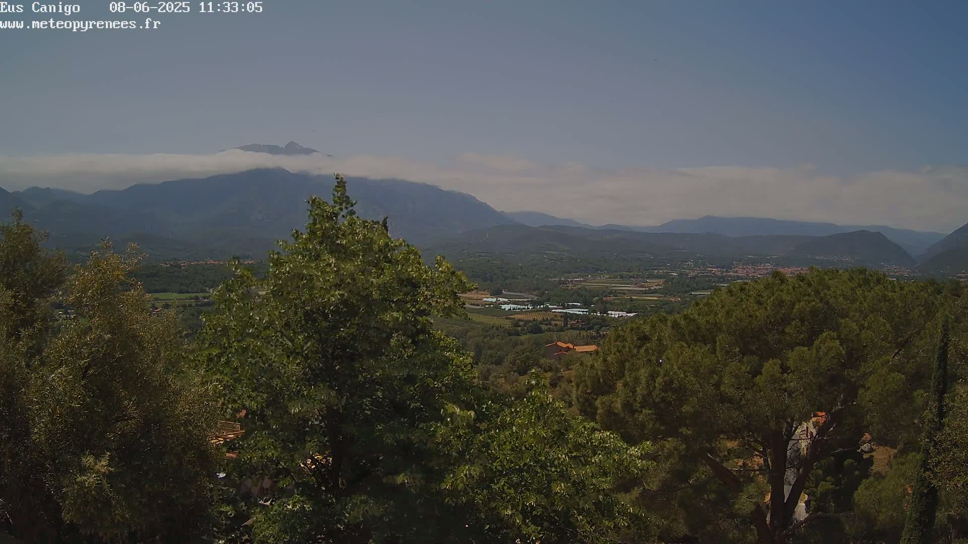 A partly cloudy day reveals a mountainous landscape viewed from a high vantage point, showing lush green vegetation and a valley stretching towards the distant mountains.