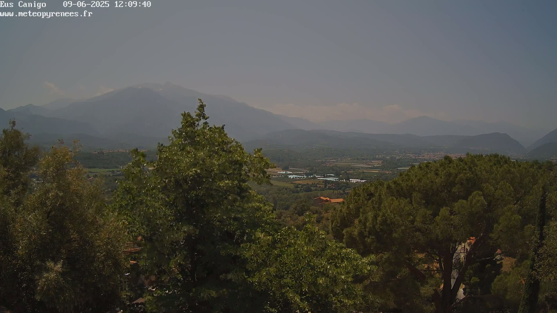 A hazy, mountainous landscape is visible from a vantage point nestled amongst green trees, showing a valley with buildings in the distance.