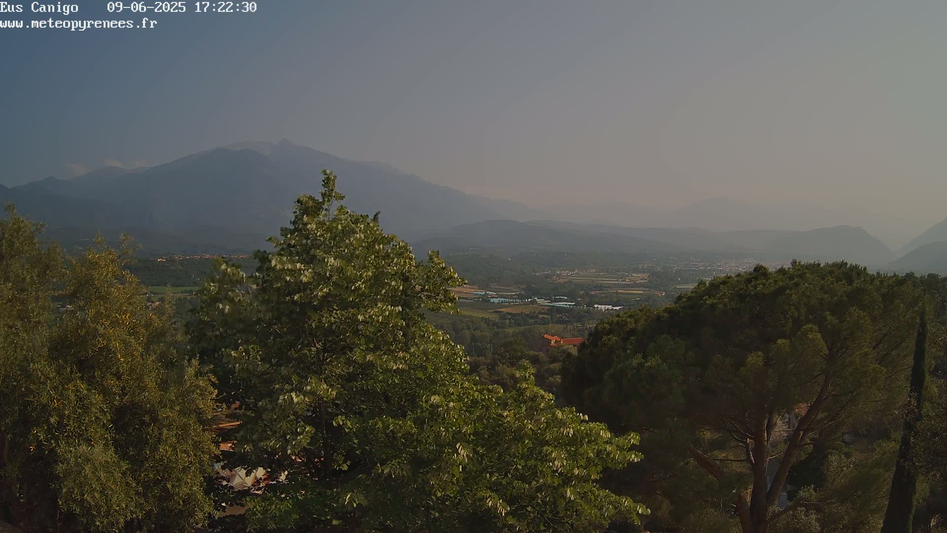 A hazy, mountainous landscape is visible from a high vantage point, showing a valley with green vegetation and distant buildings under a clear sky.