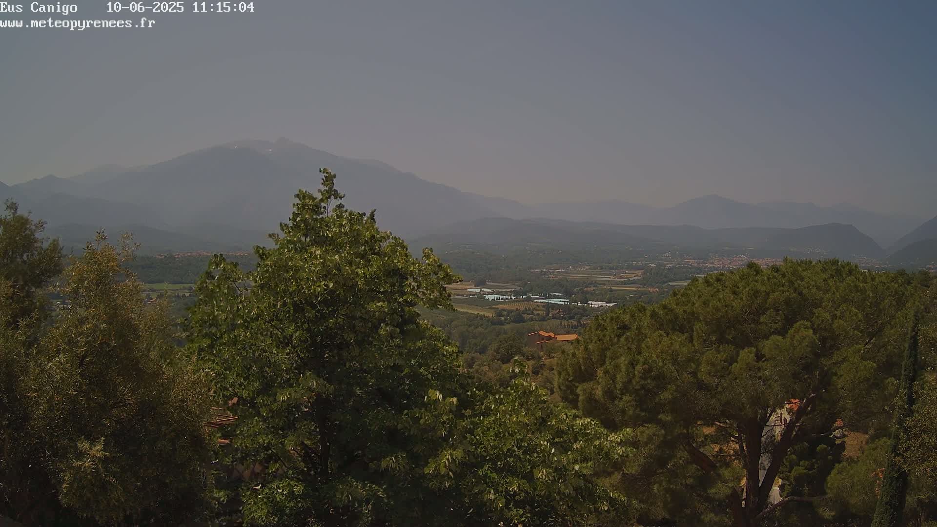 A hazy, sunny view of a mountainous landscape, seen from a high vantage point amongst lush green trees.
