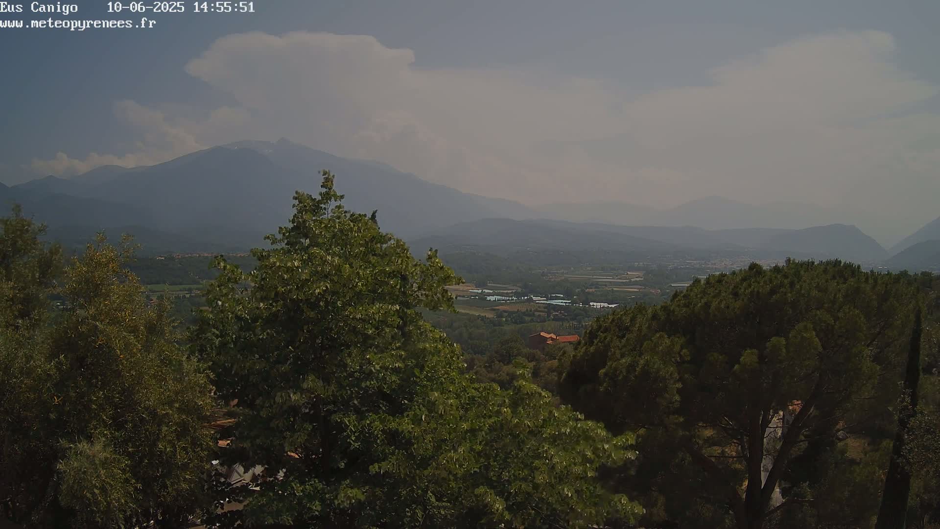 A hazy view of a mountainous landscape under a partly cloudy sky shows a valley with green trees and a few buildings visible in the distance.
