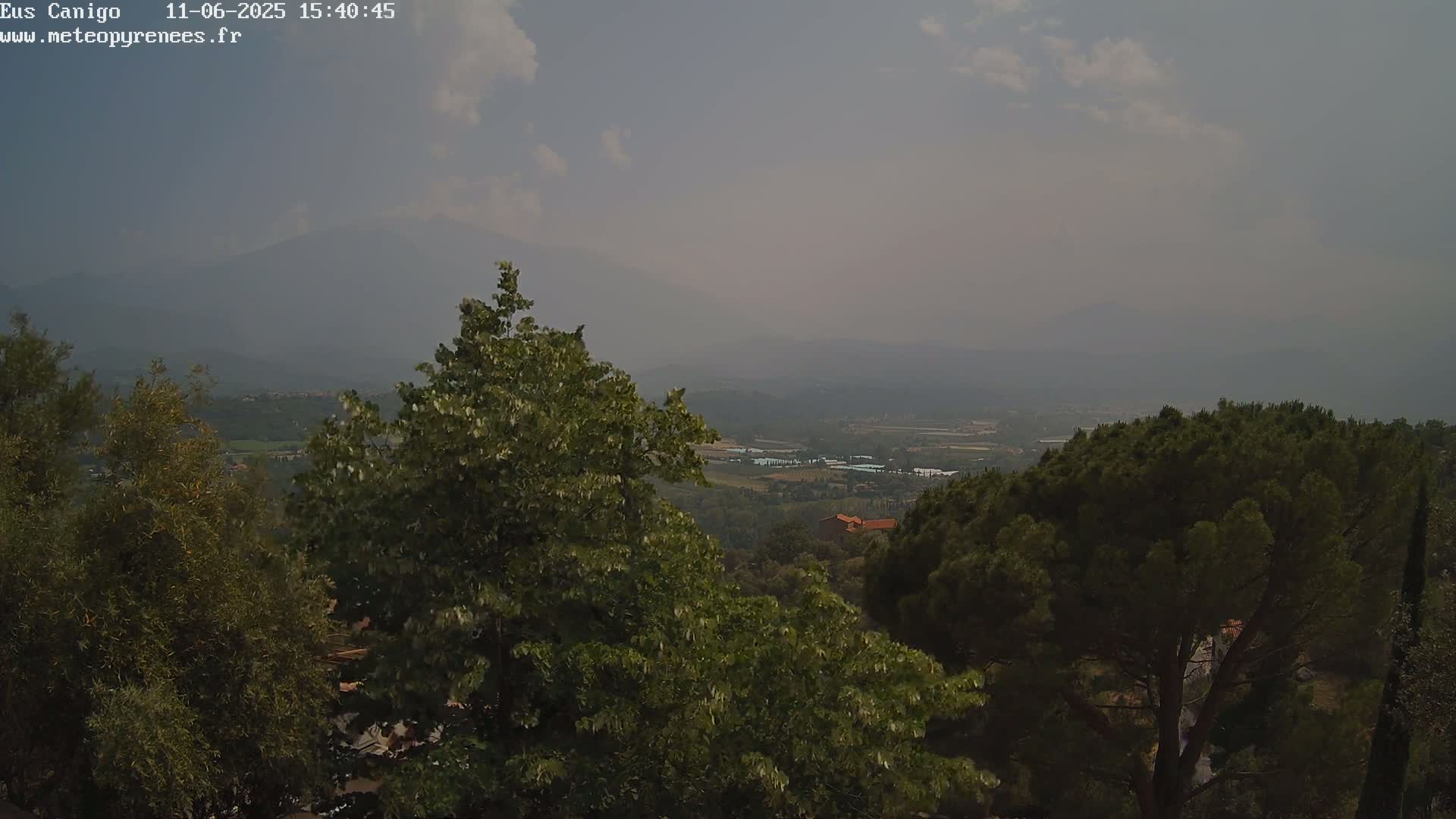 A hazy, partly cloudy view from a hilltop shows a valley with distant mountains and various trees in the foreground.