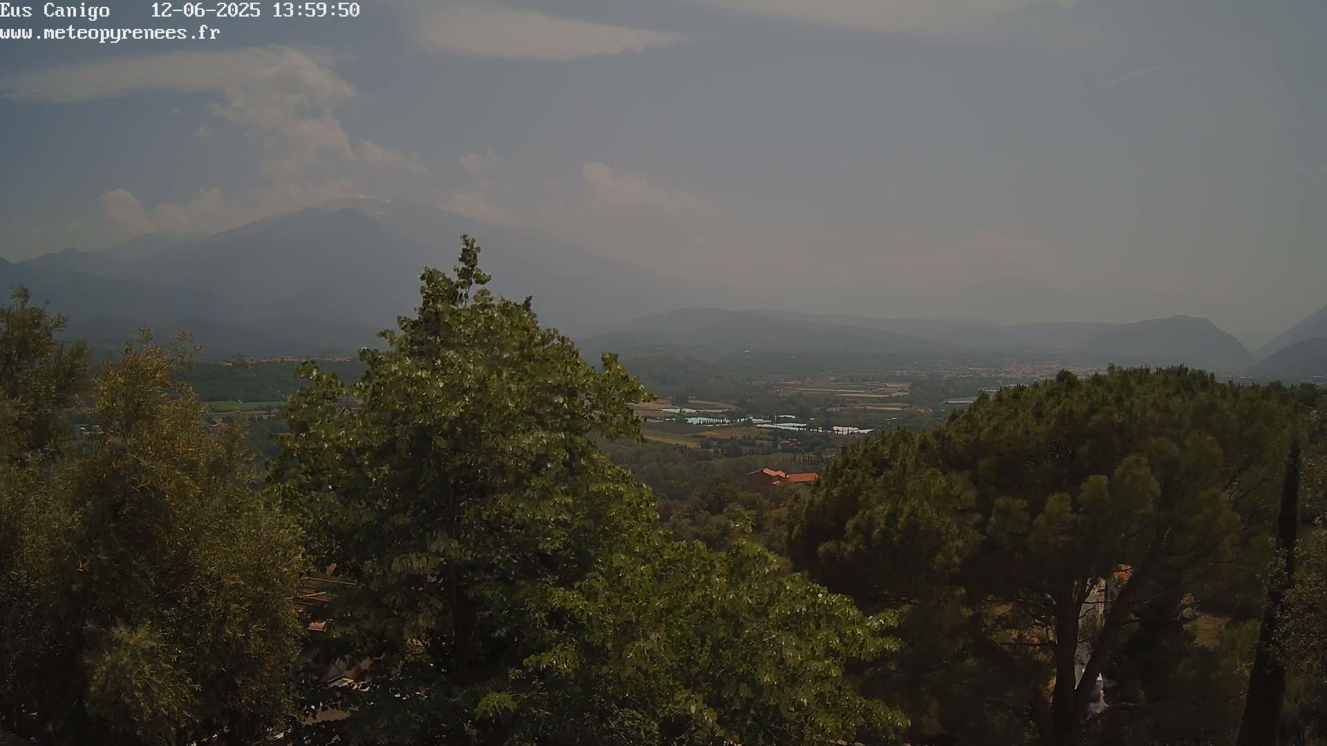 A hazy view from a hilltop shows a valley with distant mountains under a partly cloudy sky.