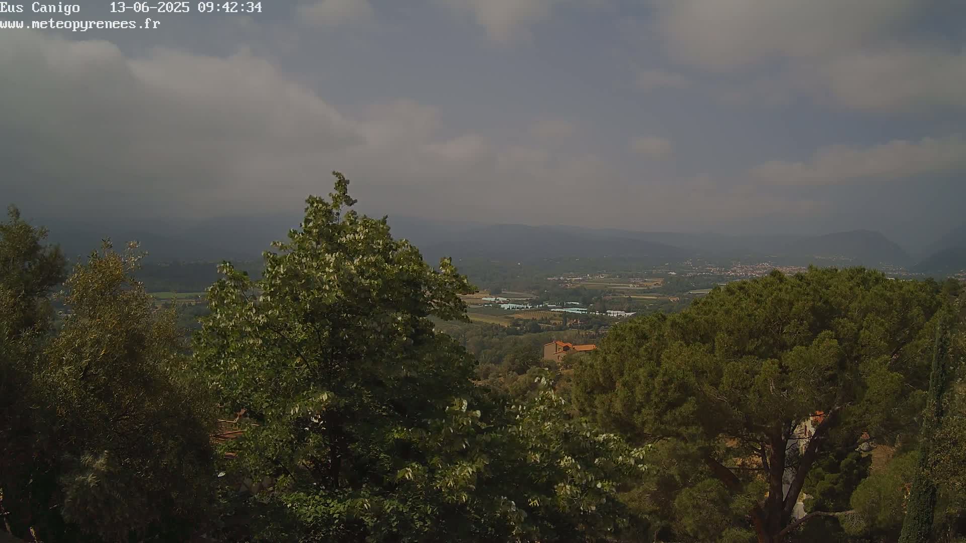 A hazy, partly cloudy day reveals a view of a valley and distant mountains seen from a high vantage point amongst green trees.