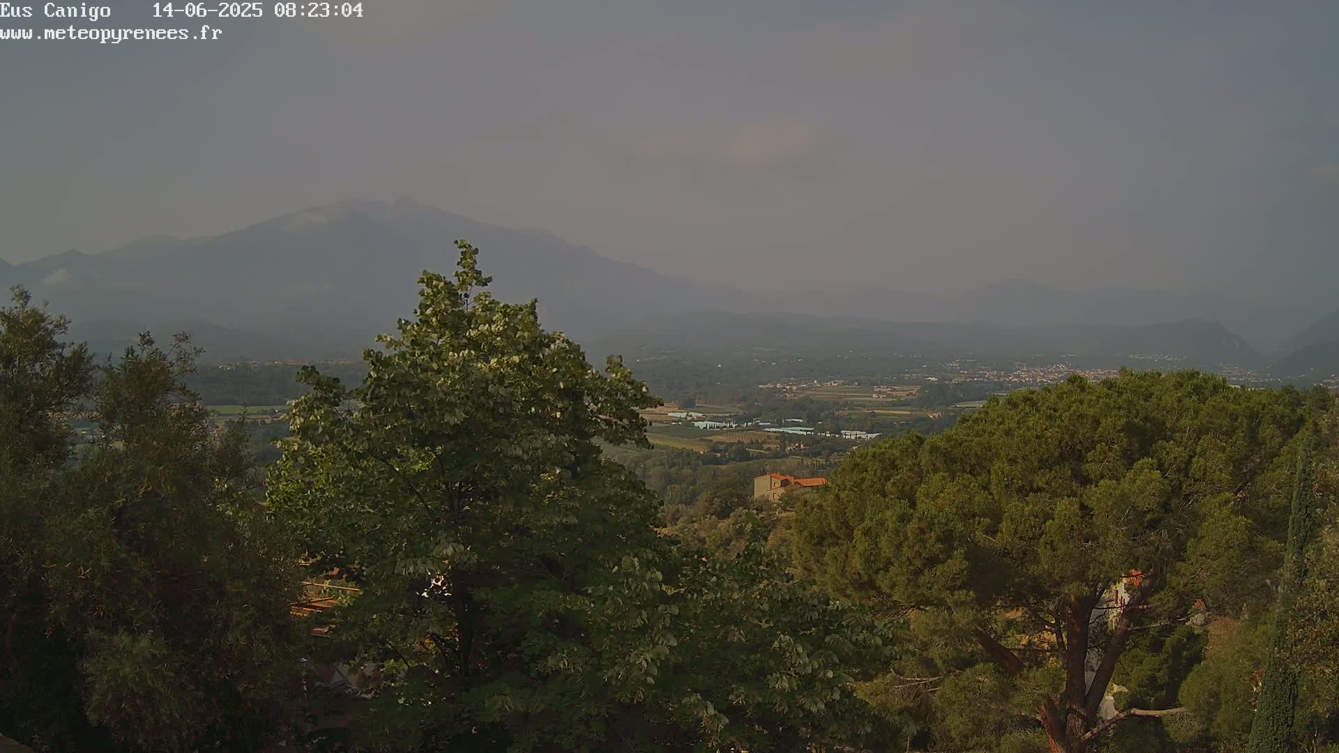 A hazy view from an elevated vantage point shows a valley dotted with buildings and greenery, with mountains in the background and lush green trees in the foreground.