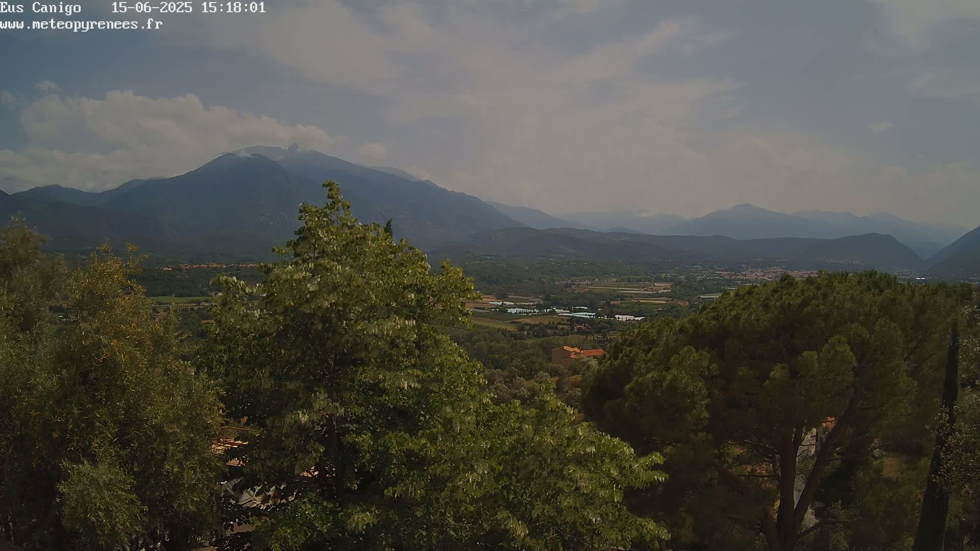A hazy, mountainous landscape is visible from an elevated viewpoint, with lush green trees in the foreground and a valley extending to distant ranges under a partly cloudy sky.