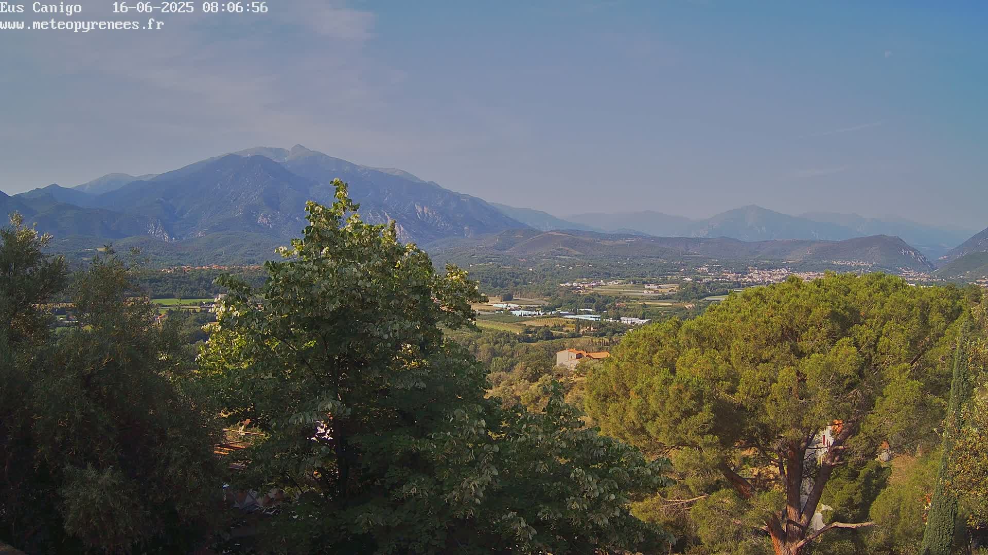 A mountainous landscape under a clear, light-blue sky is viewed from a slightly elevated vantage point, showing lush green trees in the foreground and a valley with scattered buildings in the mid-ground.