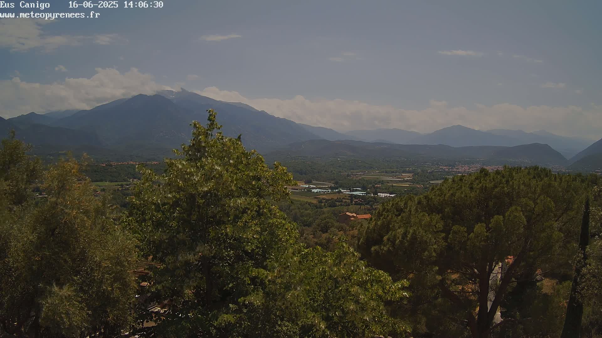 A partly cloudy sky overlooks a mountainous landscape dotted with green trees and vegetation, revealing a valley with buildings in the distance.