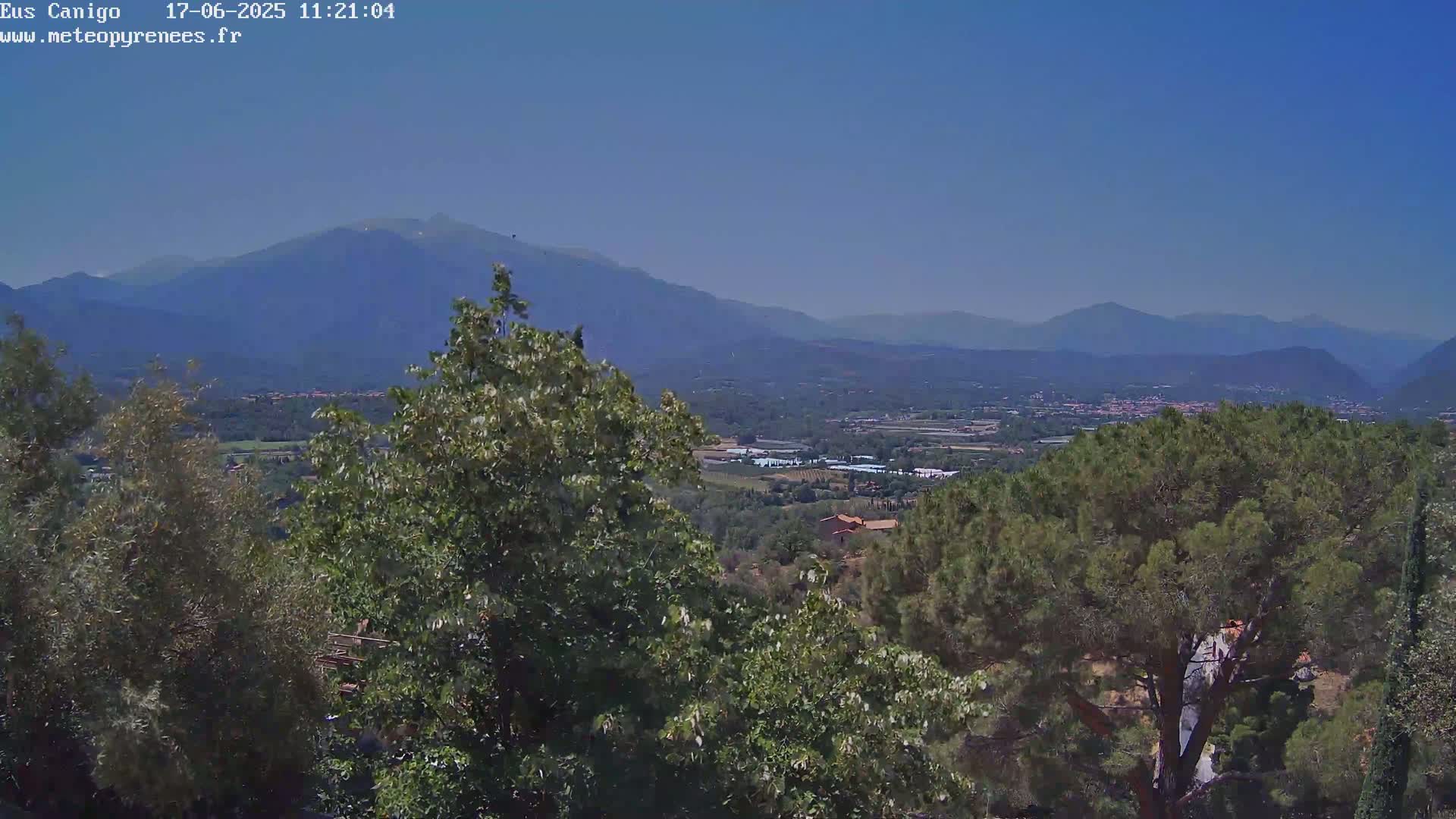 A clear, sunny day reveals a panoramic view of a valley nestled between distant mountains, partially obscured by lush green trees in the foreground.
