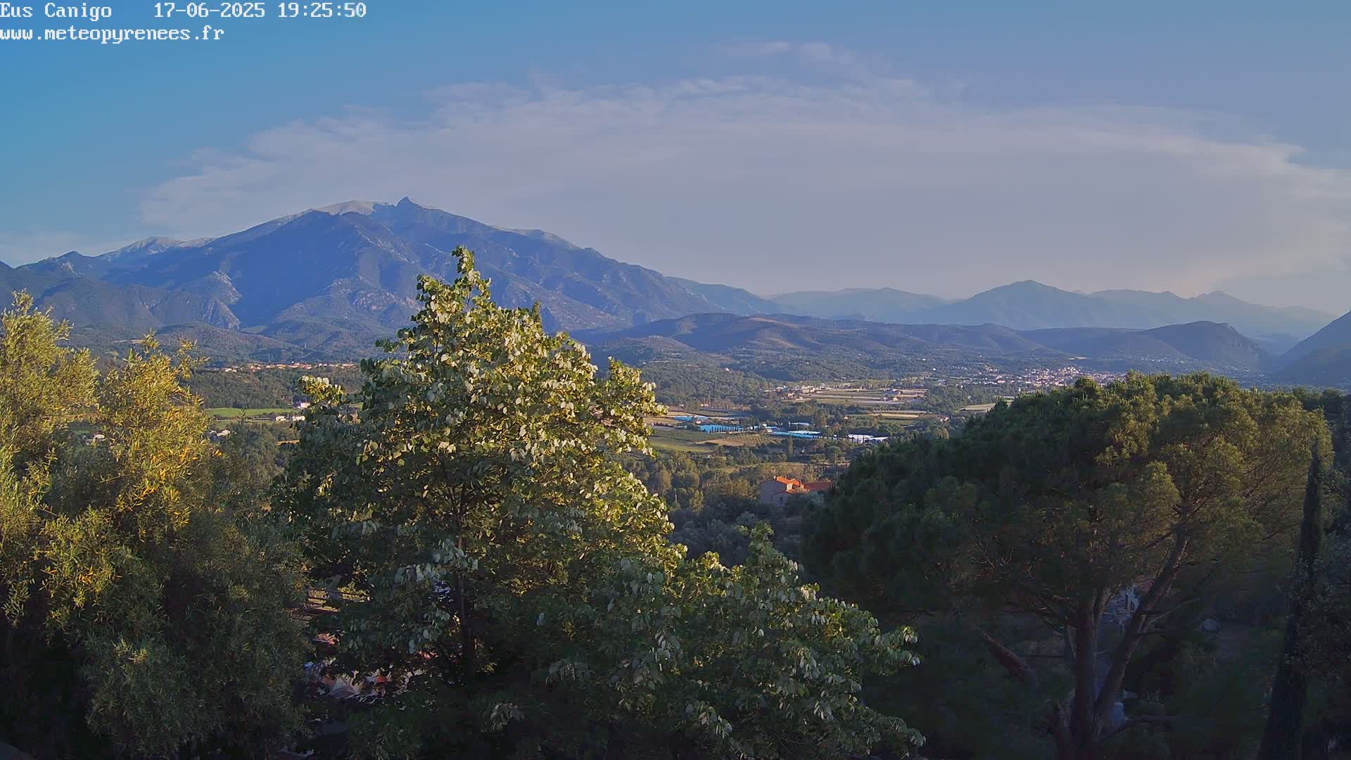 A mountainous landscape under a mostly clear sky shows a valley with trees and buildings in the foreground, leading to a range of distant mountains.