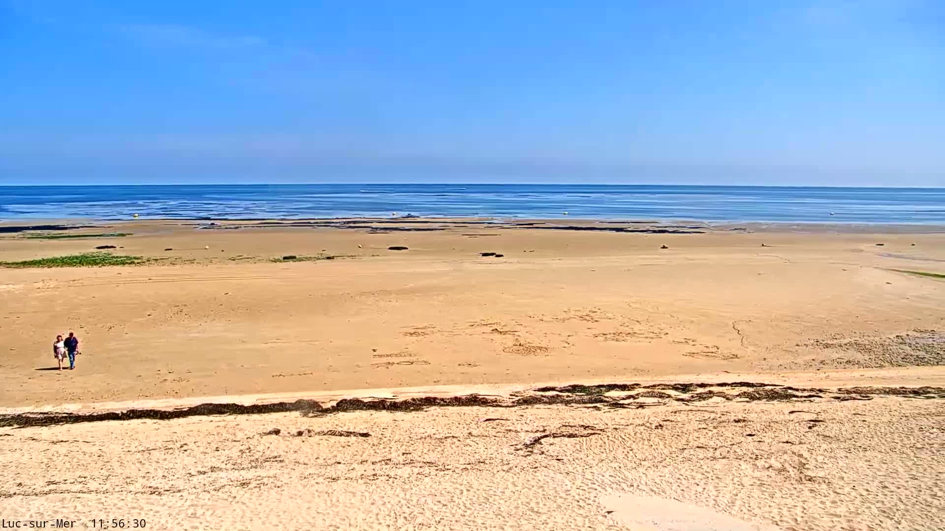 A wide, sandy beach stretches to a calm, blue ocean under a clear sky, with two people walking on the sand in the distance.