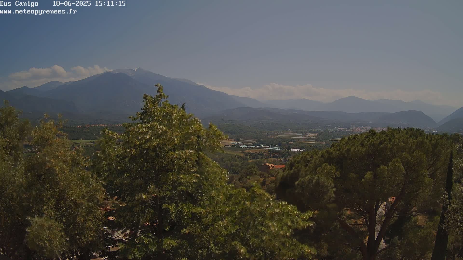 A partly cloudy day reveals a mountainous landscape viewed from a vantage point amongst lush green trees, showing a valley with fields and buildings in the middle ground.