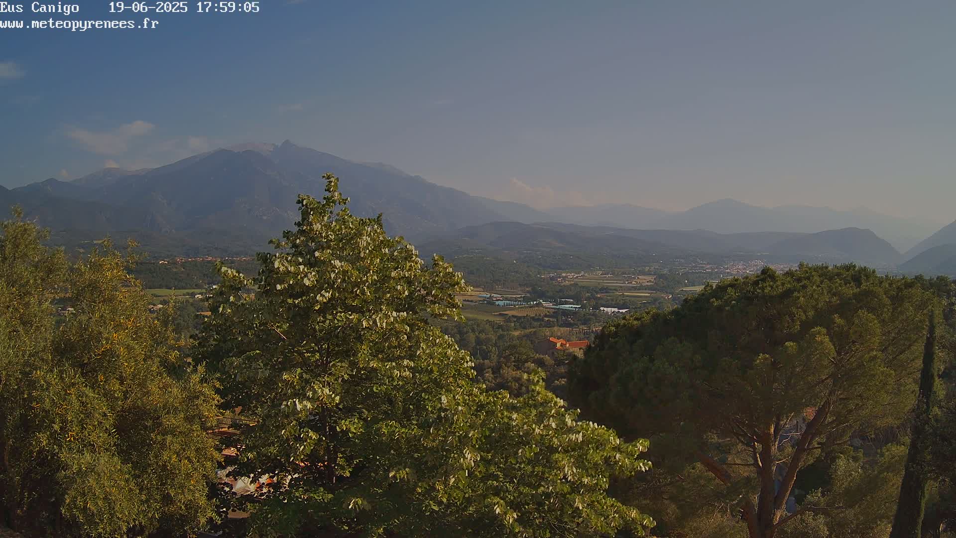 A mountain range is visible in the distance under a clear sky, with a valley of trees and buildings in the foreground.