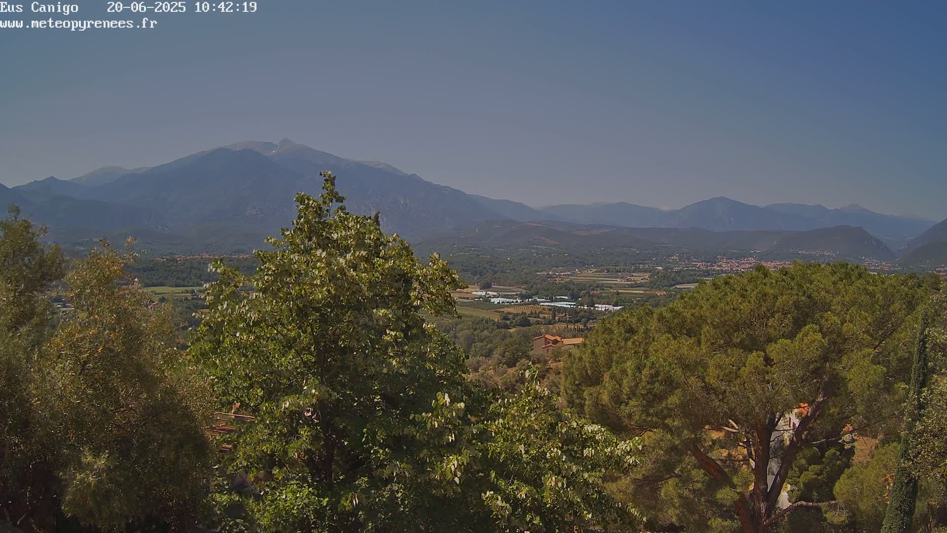 A panoramic view of a valley and distant mountains under a clear sky, partially obscured by lush green trees in the foreground.