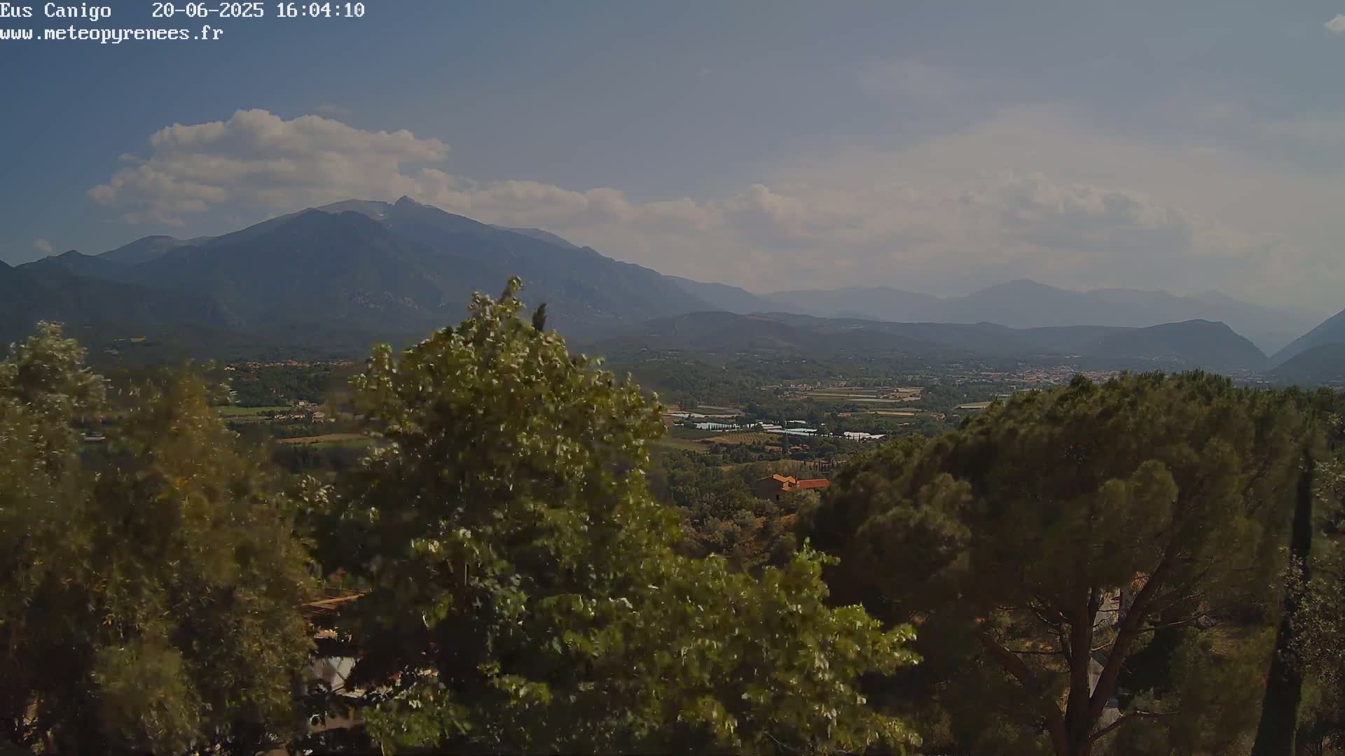 A partly cloudy day reveals a mountainous landscape, with a valley of greenery and small buildings nestled between rolling hills and distant peaks.