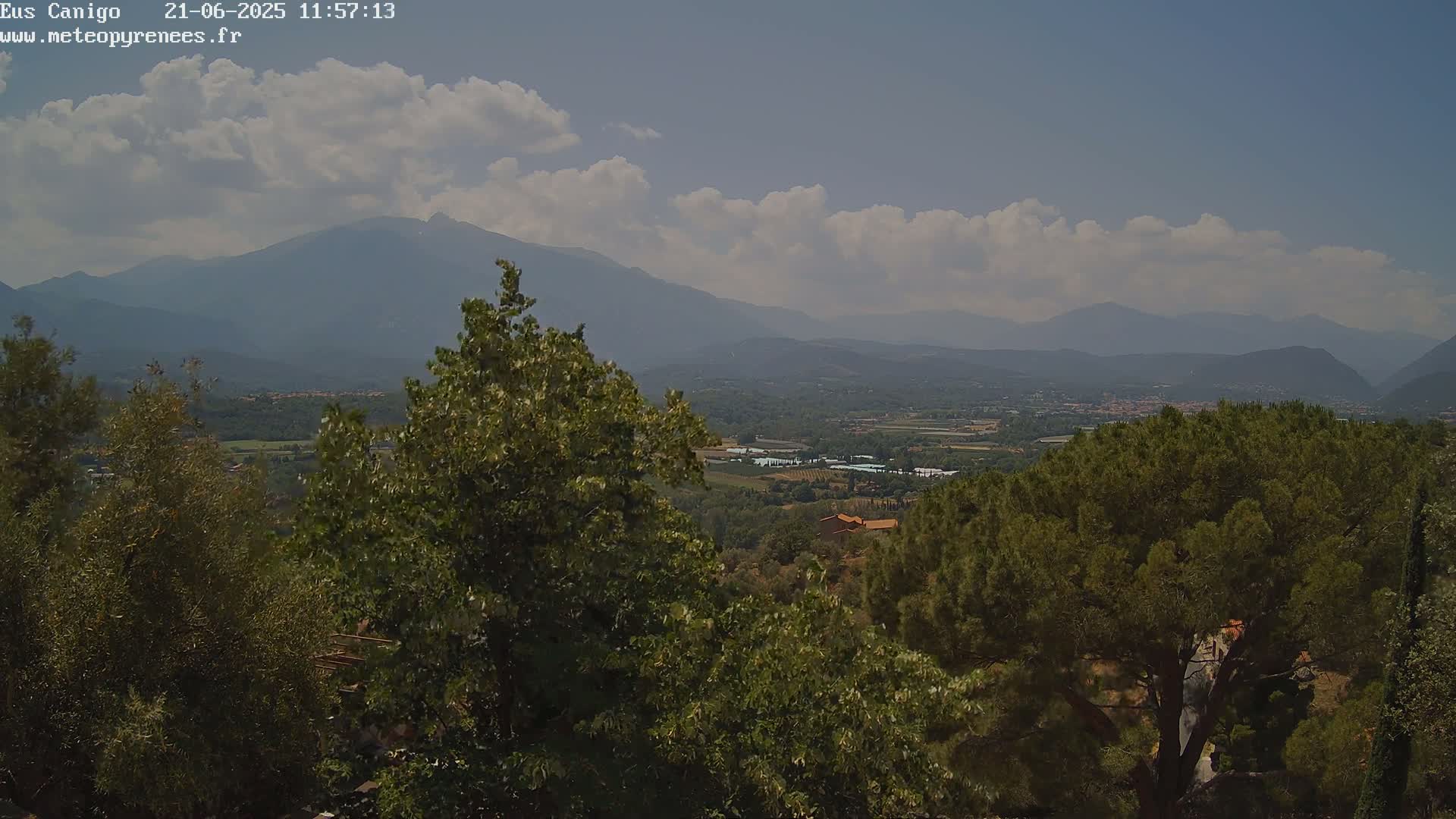 A partly cloudy day reveals a mountainous landscape viewed from a high vantage point, showing a valley with greenery and some structures nestled between hills and mountains in the distance.