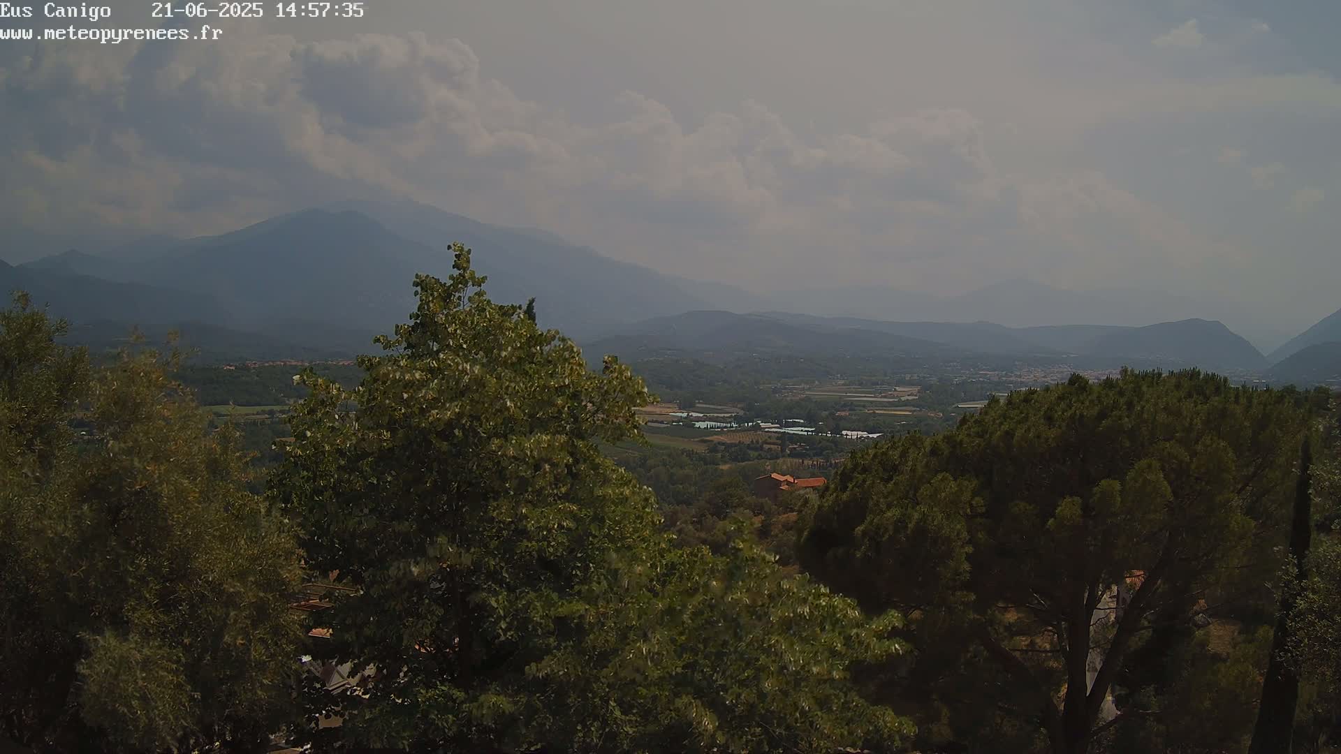 A hazy, partly cloudy day reveals a mountainous landscape with a valley filled with vegetation and buildings visible in the distance, observed from a vantage point amongst leafy trees.