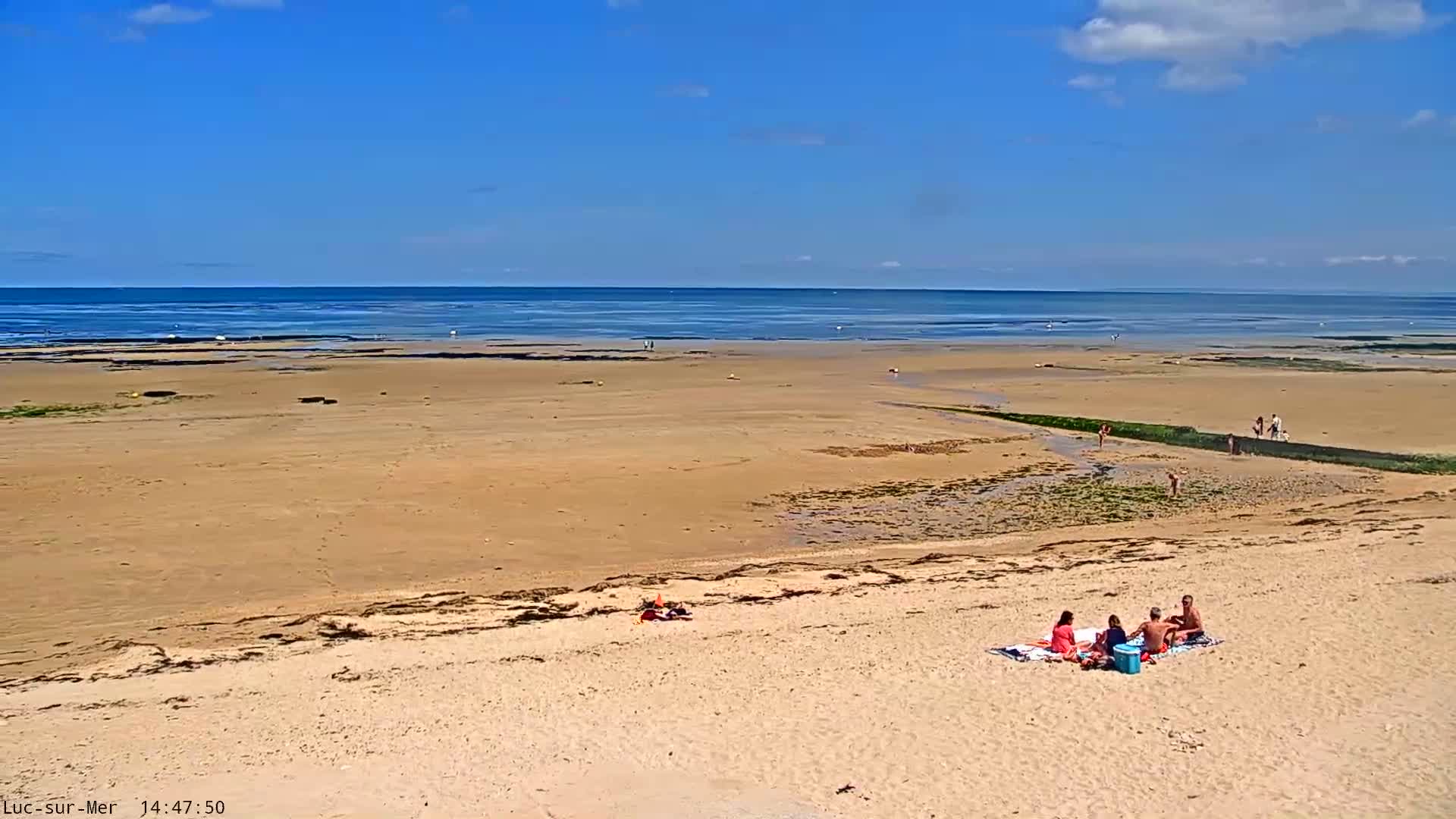 A wide sandy beach stretches to a calm ocean under a mostly sunny sky, with a small group of people sitting on a blanket near the top of the beach.