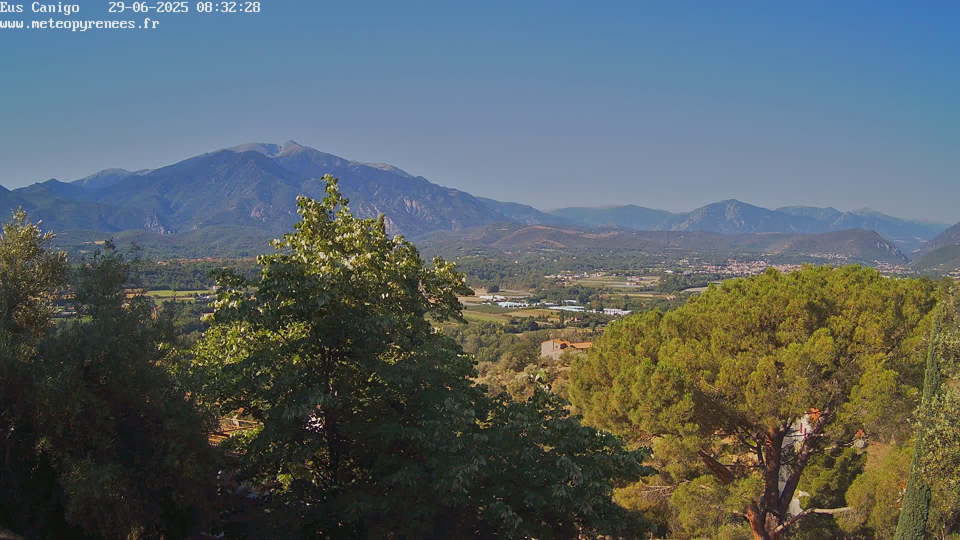 A mountain range stretches across the horizon under a clear blue sky, viewed from a hillside covered in lush green trees and shrubs.