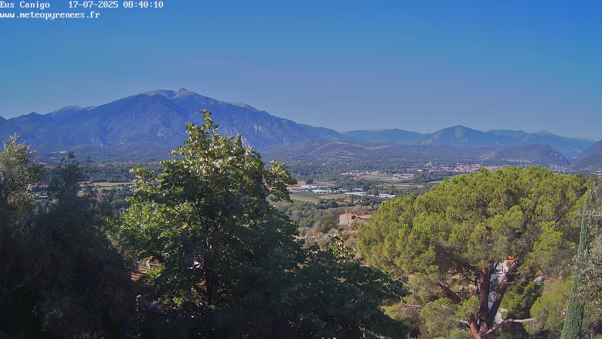 A mountain range stretches across a clear blue sky, overlooking a valley of green trees and small buildings.