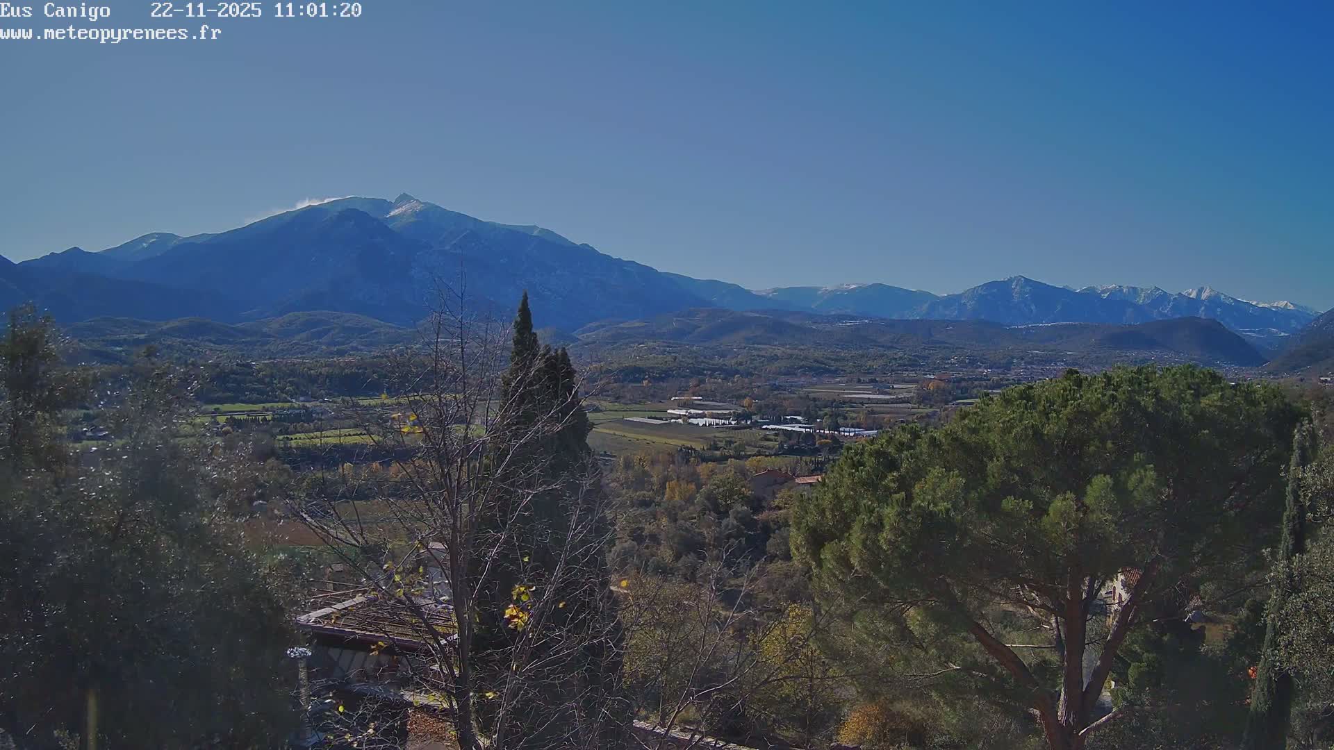 A vast outdoor landscape on a clear, sunny day presents a valley dotted with towns and agricultural fields displaying autumn colors, framed by a mix of bare and evergreen trees in the foreground, with majestic, snow-capped mountains rising in the distant background under a brilliant blue sky.