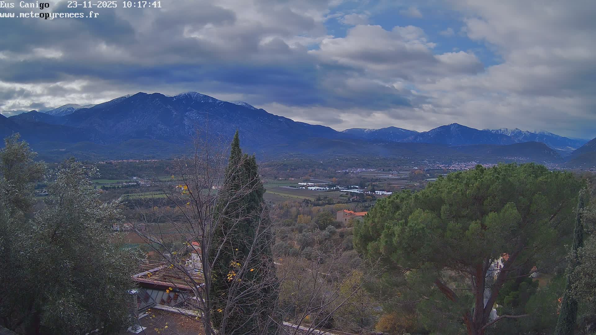 On a partly cloudy day, a valley containing fields and scattered villages is visible, framed by various trees in the foreground and backed by dark, snow-capped mountains.
