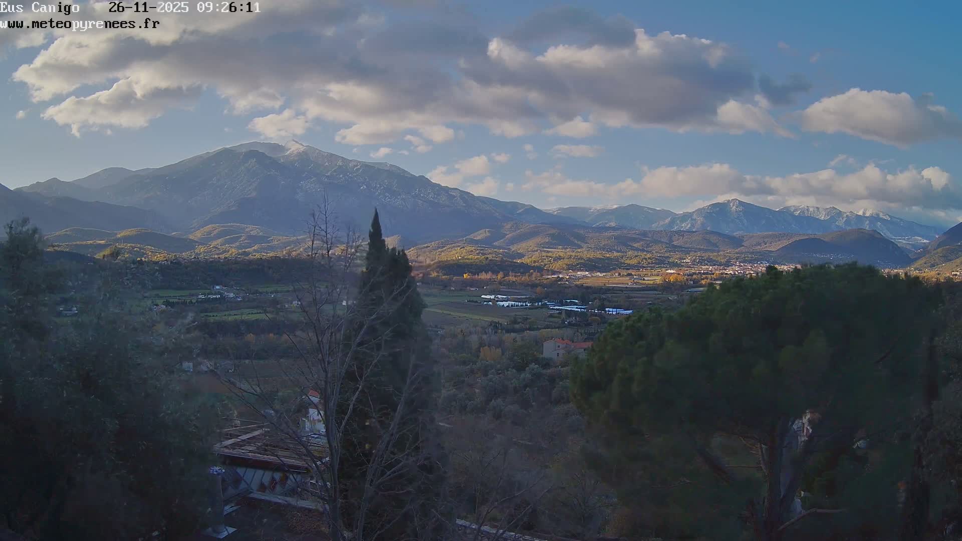 A panoramic view reveals a sun-drenched valley filled with fields and scattered settlements, framed by forested hills and majestic, snow-capped mountains under a partly cloudy blue sky.