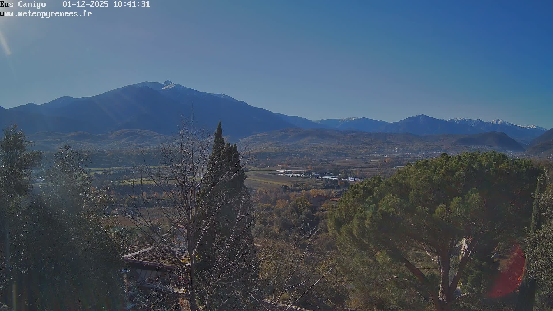 A clear blue sky on a sunny day overlooks a wide valley with autumnal trees and scattered buildings, framed by foreground foliage, leading to a range of mountains, some with snow-capped peaks, in the distance.