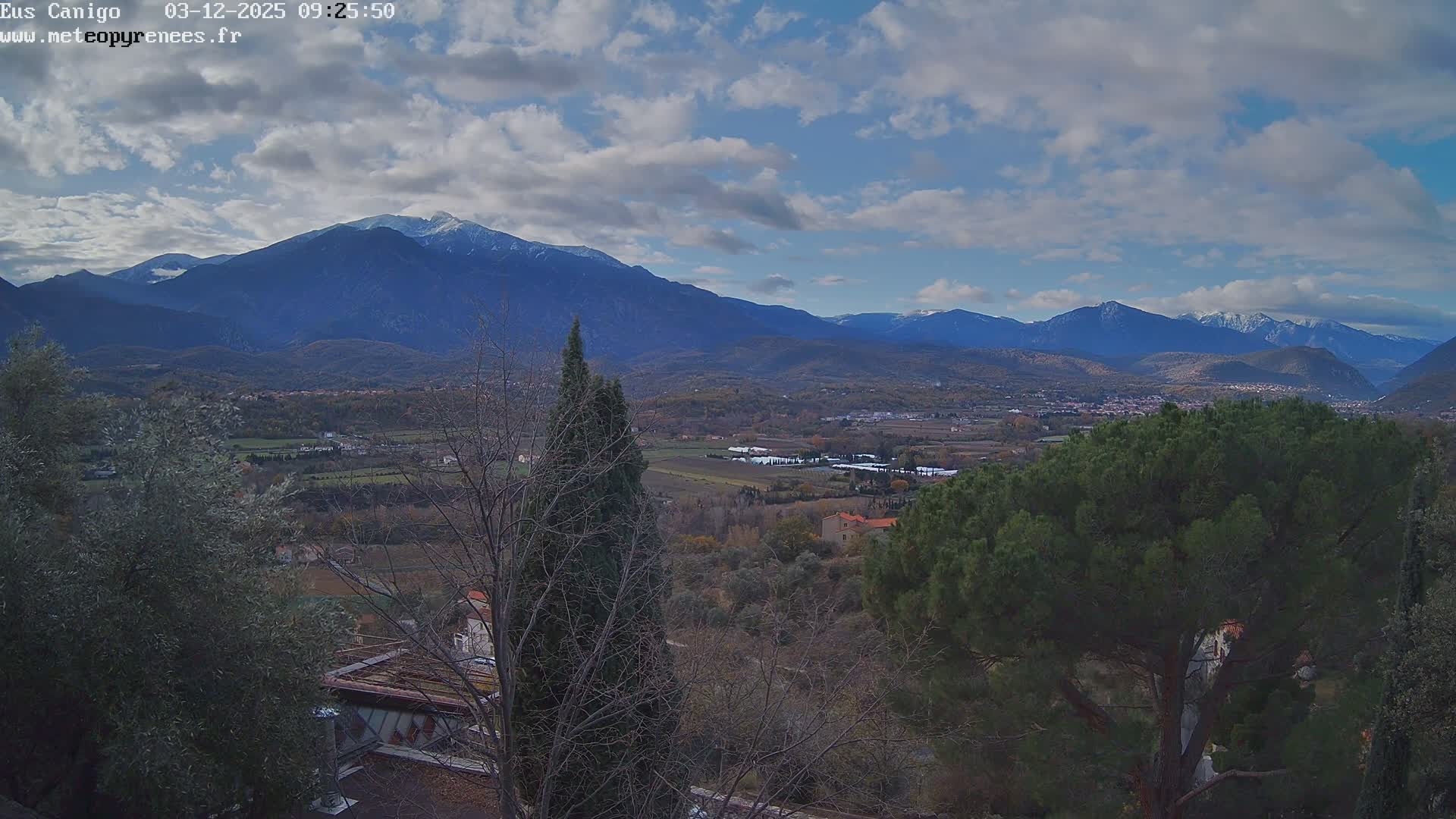 A vast valley landscape is captured under a partly cloudy sky, featuring a mix of evergreen and bare trees in the foreground, leading to a patchwork of fields and scattered villages, with a majestic range of distant, snow-capped mountains dominating the background.