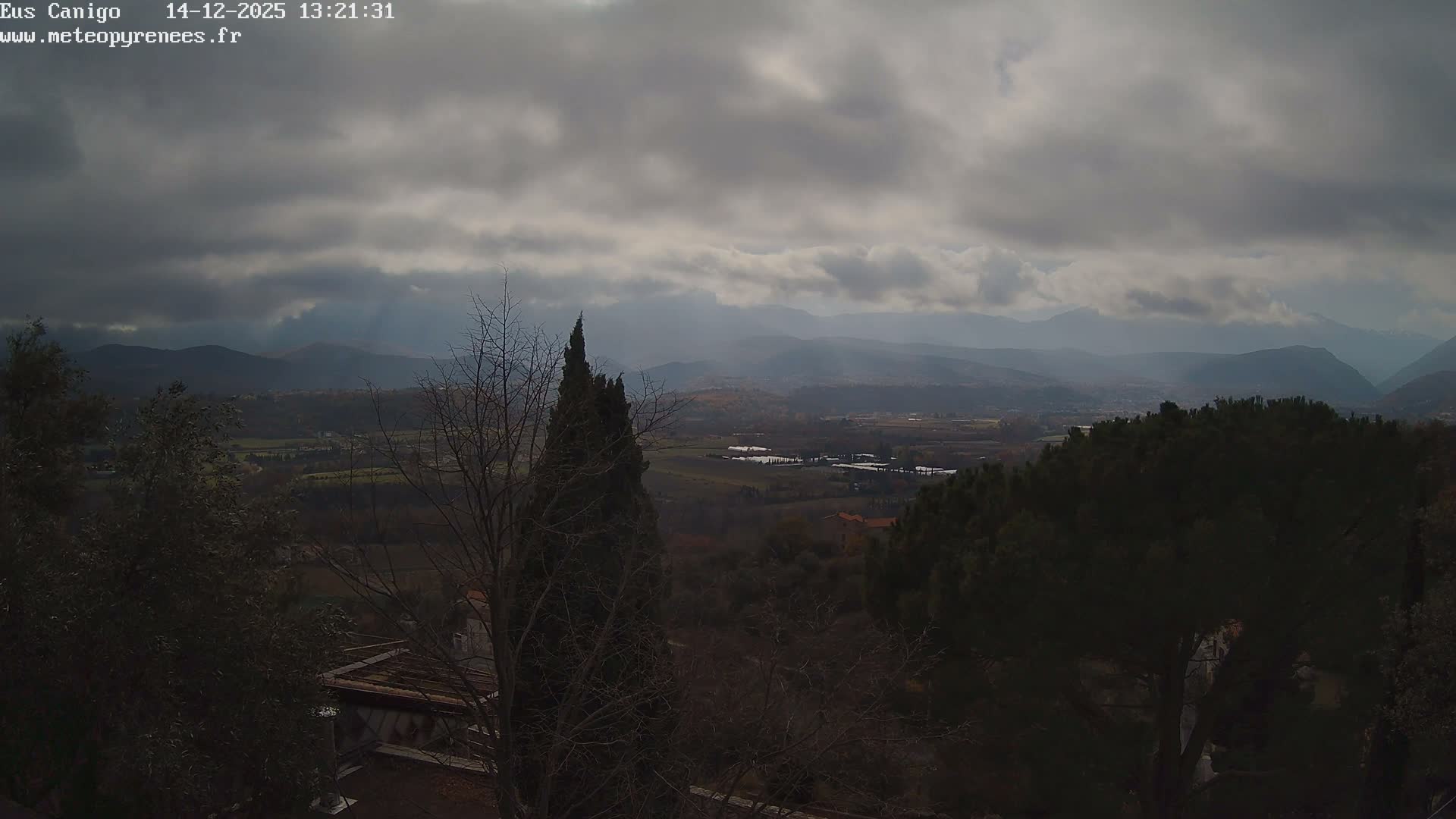 A vast valley landscape is captured under a partly cloudy sky, featuring a mix of evergreen and bare trees in the foreground, leading to a patchwork of fields and scattered villages, with a majestic range of distant, snow-capped mountains dominating the background.