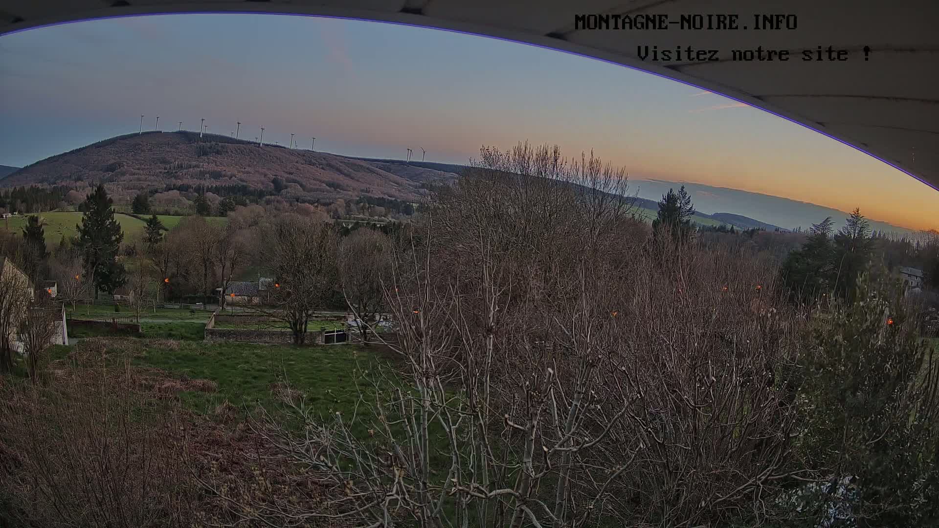 A sunset view of a hilltop wind farm, overlooking a rural landscape with bare trees and patchy green fields under a clear sky.