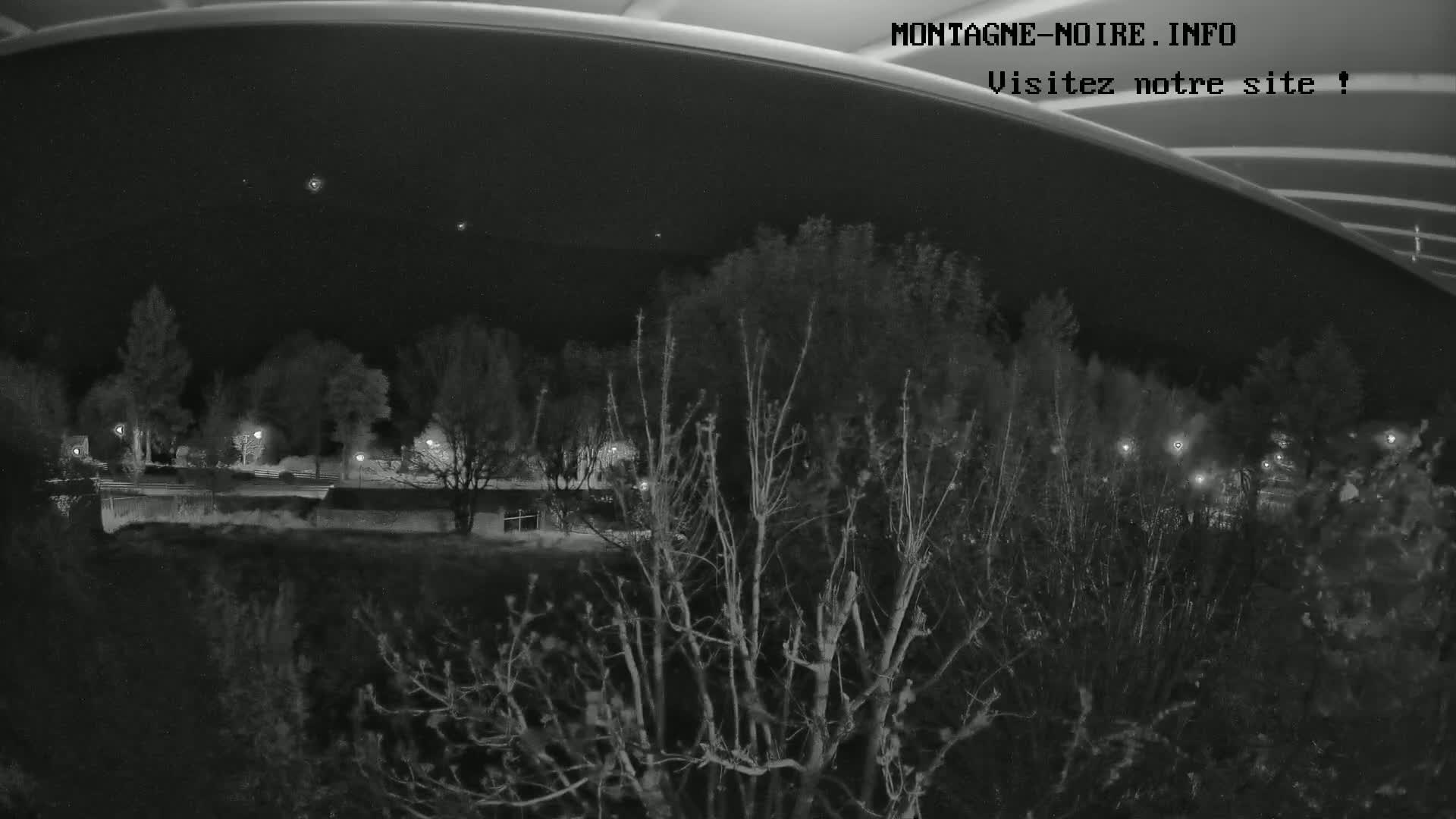 A nighttime view from under a bridge shows bare trees and dimly lit buildings across a body of water.