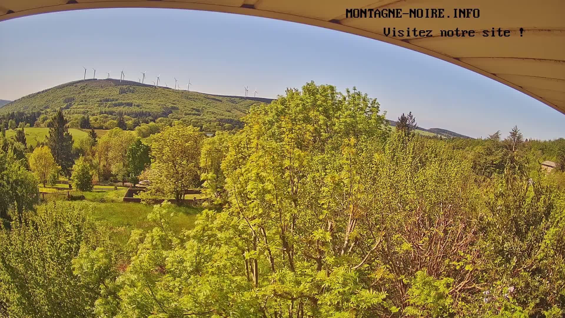 A view from a partially covered vantage point shows a verdant landscape under a clear blue sky, with a hillside dotted with wind turbines in the distance.