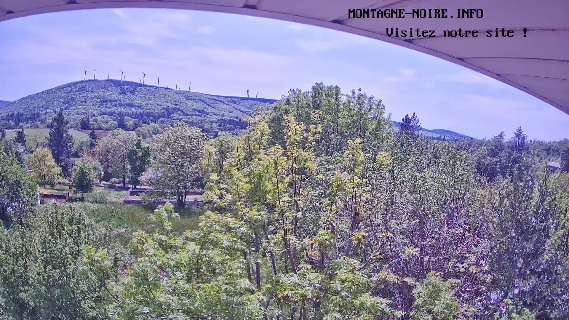 A view from under a curved awning shows a sunny day with a lush green landscape dotted with trees, a distant hill topped with wind turbines, and a clear blue sky.