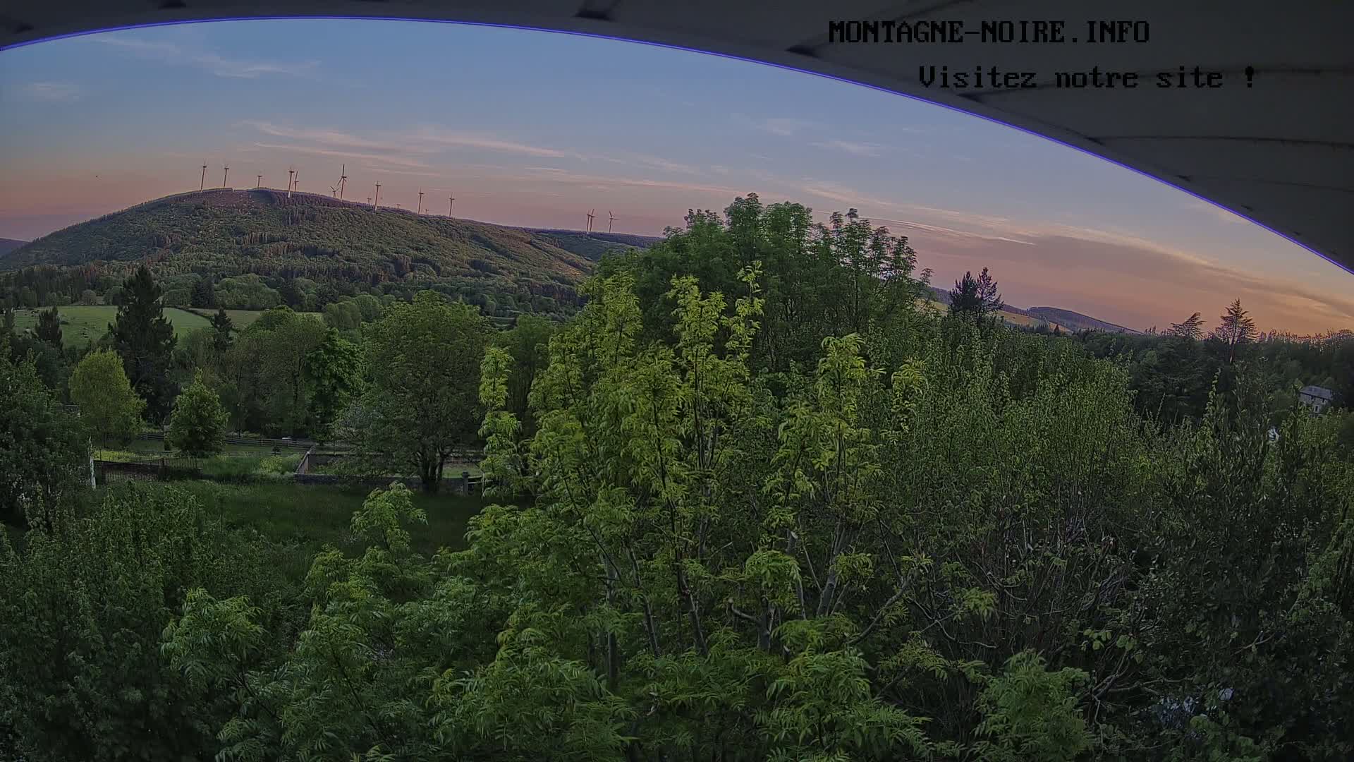A view from an elevated position shows a lush green landscape at sunset, with a hilltop dotted with wind turbines in the distance.