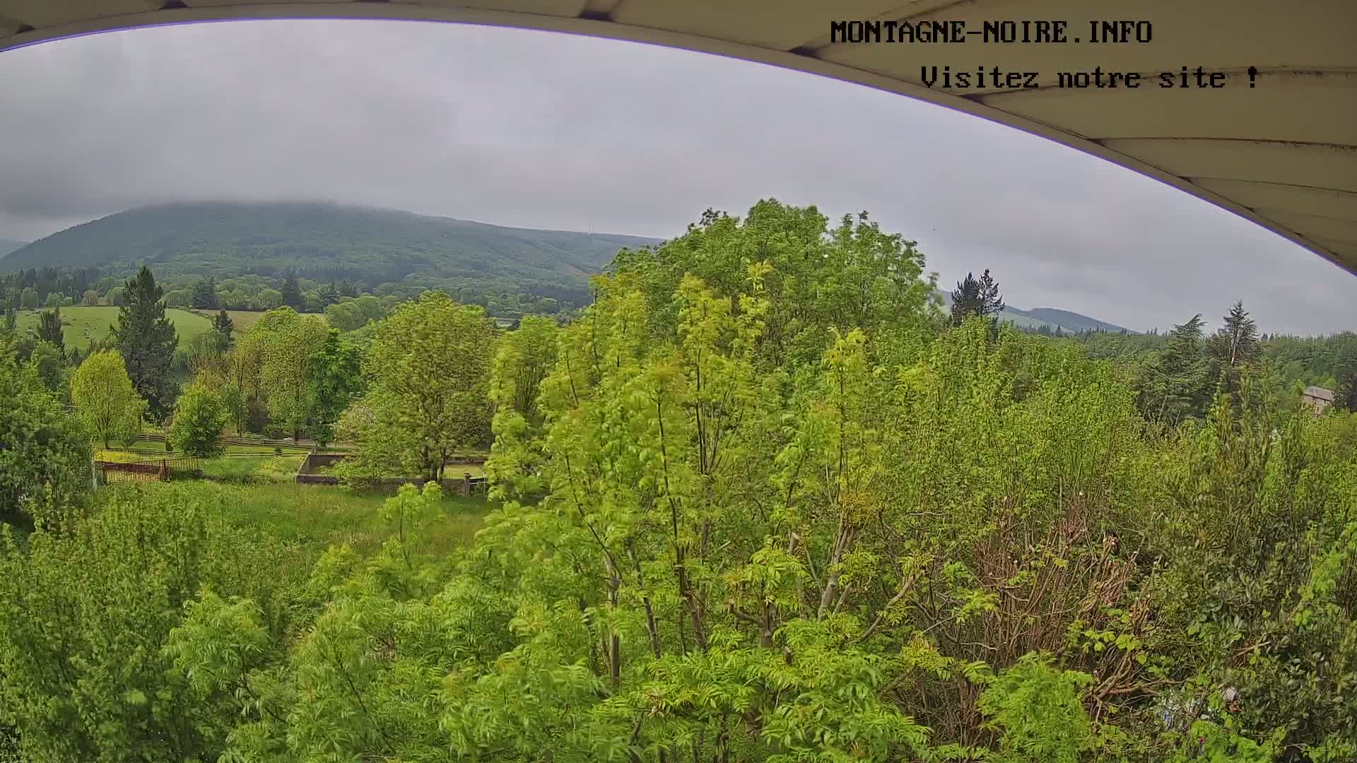 A view from under a covered structure shows a lush green landscape with a hazy mountain in the background under an overcast sky.