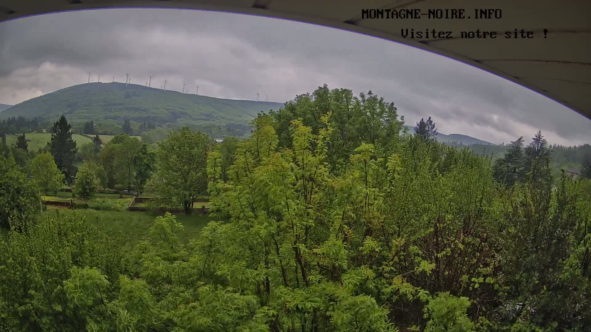 A view from under a covered structure shows a lush green landscape with a distant hill topped with wind turbines under an overcast sky.