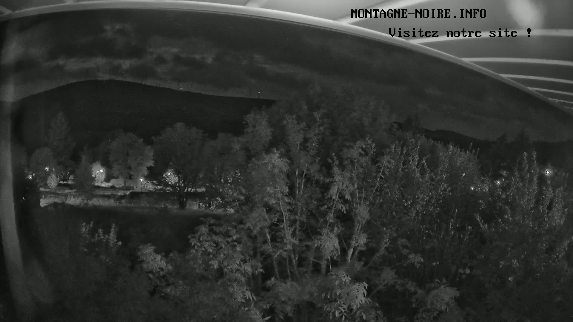 A nighttime view from behind a curved surface shows a dark hill in the background, with vegetation and faintly lit roads in the foreground under a cloudy sky.