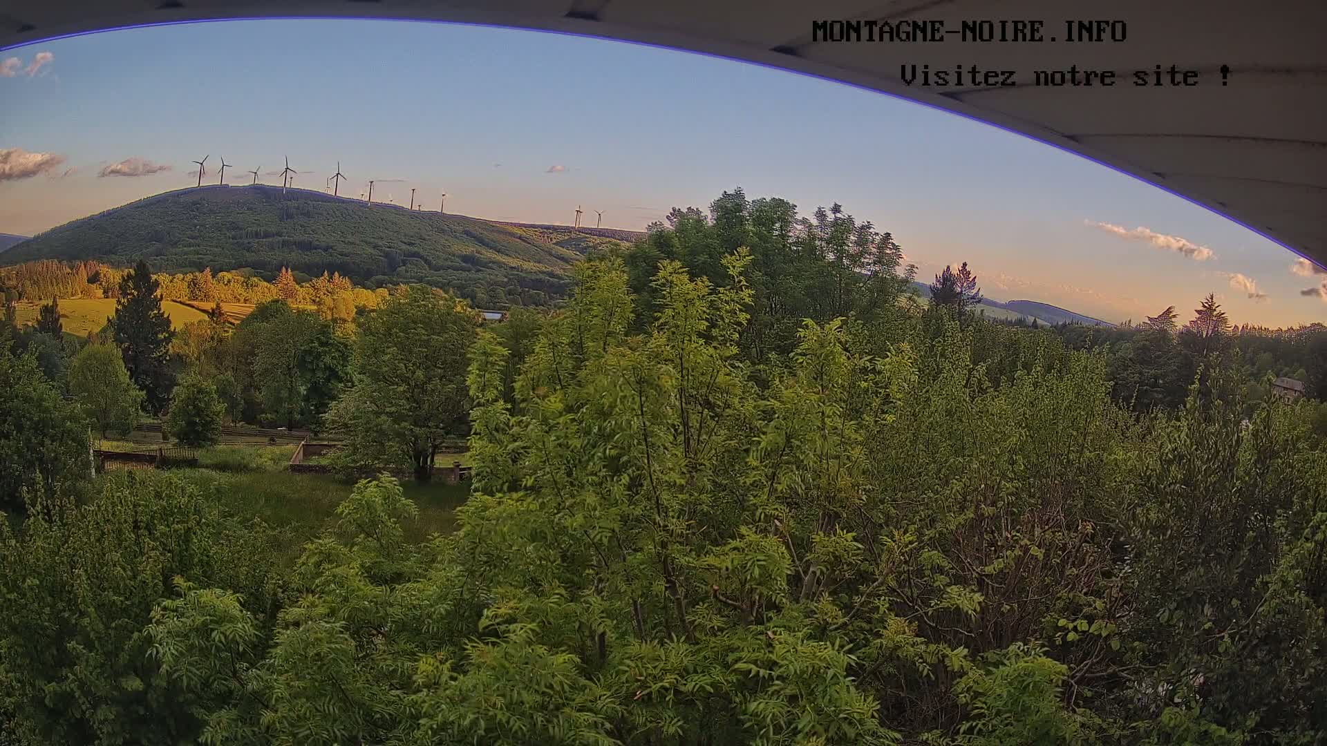 A view from an elevated position shows a lush green landscape with a hill in the background topped with wind turbines under a partly cloudy, twilight sky.
