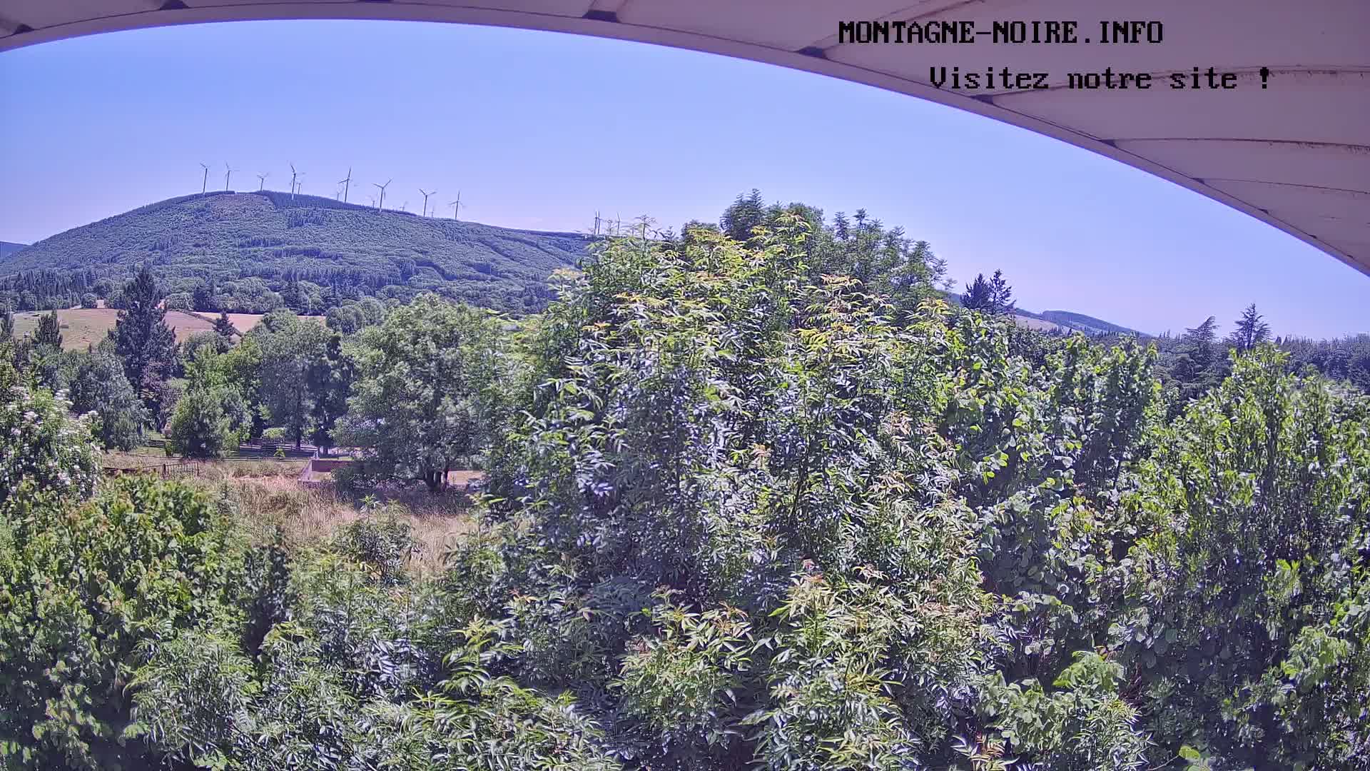A view from under an awning shows a lush green landscape with a distant hill topped with wind turbines under a clear blue sky.