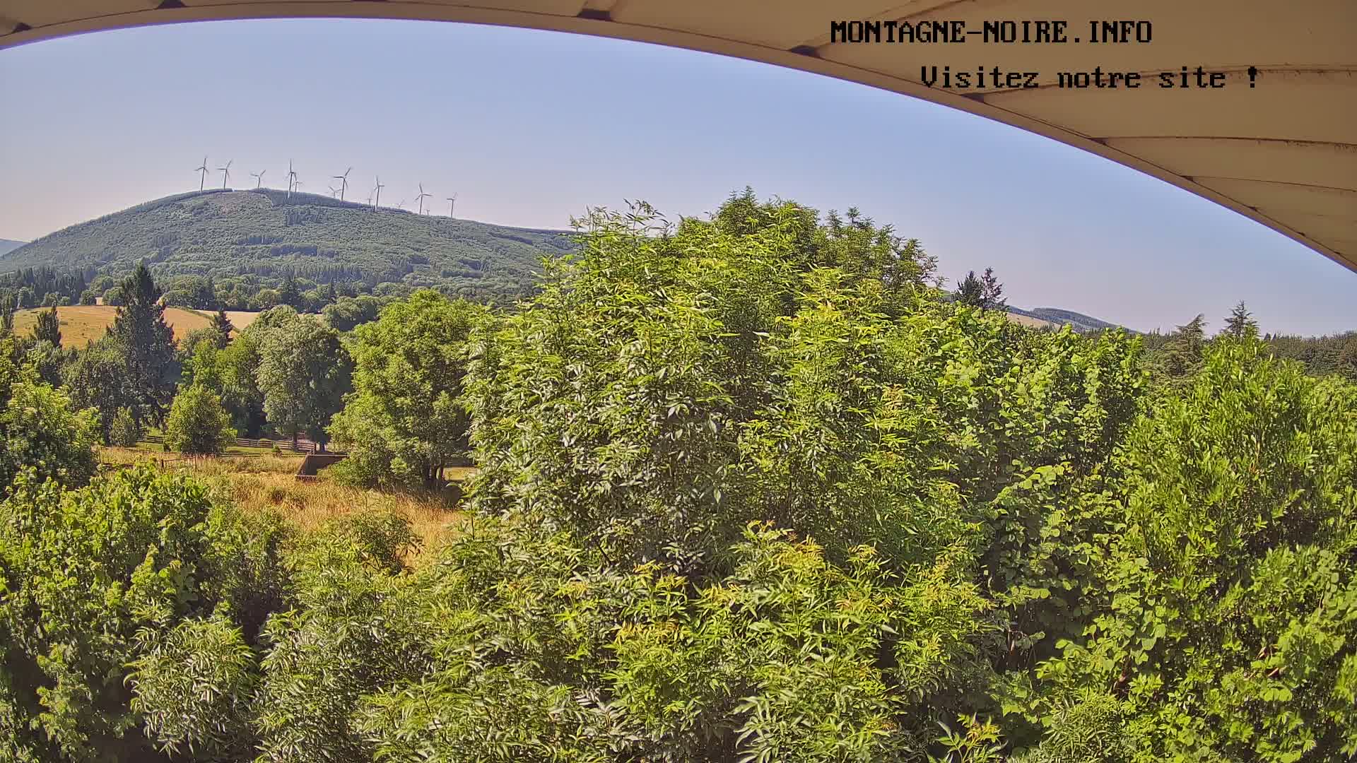 A sunny view from a partially covered vantage point shows a verdant landscape with a distant hill topped by wind turbines and lush green trees and fields in the foreground.