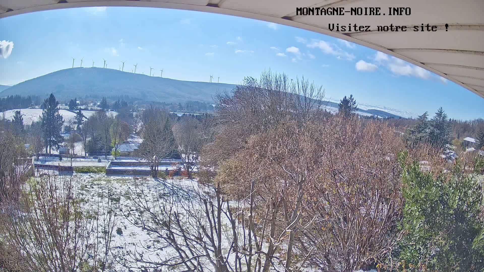 A bright, sunny winter day reveals a snow-dusted landscape of bare trees and fields in the foreground, stretching towards a valley and a distant mountain ridge adorned with wind turbines, all beneath a clear blue sky with scattered white clouds.